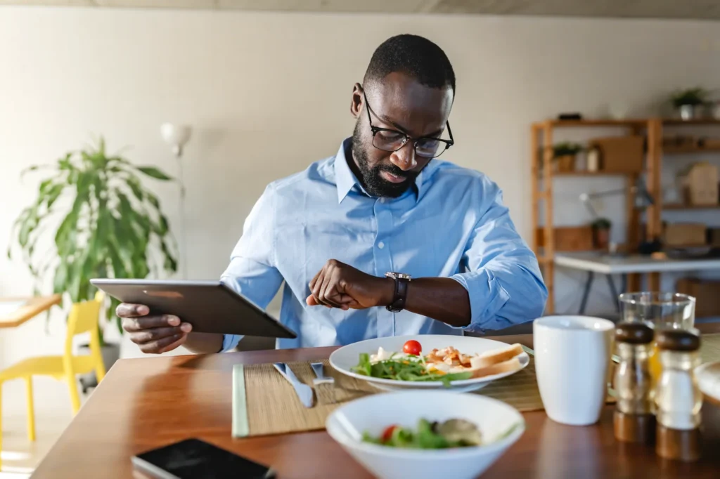 Man checking his watch while holding a tablet and eating a healthy meal.