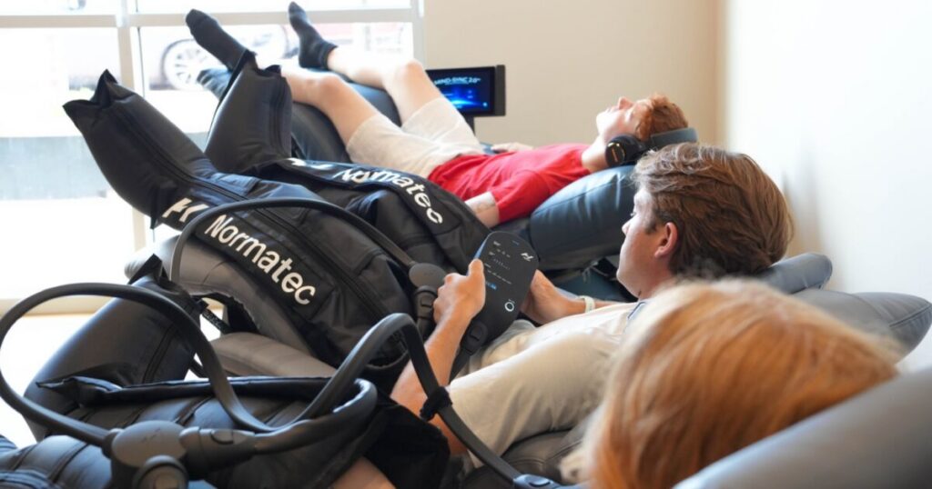 Three clients receiving Normatec compression therapy in reclining chairs at Sisu Longevity Studio in Colorado Springs, one adjusting the controller.